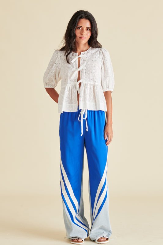 A female model wearing a white, short-sleeved top with a tie-front neckline and blue wide-leg pants with white side stripes.